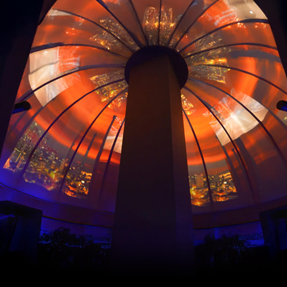 A spectacular 360-degree projection transforms a domed ceiling into a vibrant sunset sky with geometric patterns. The silhouette of a person stands centered beneath the radiant display of orange, red, and blue hues that create an otherworldly atmospheric experience.